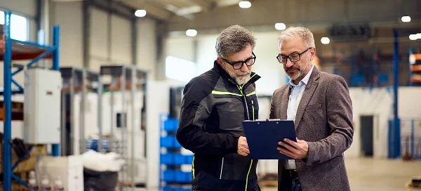 Two men standing in a warehouse discuss information on a clipboard; one wears a business suit, the other wears a work jacket. Shelving and equipment are visible in the background.
